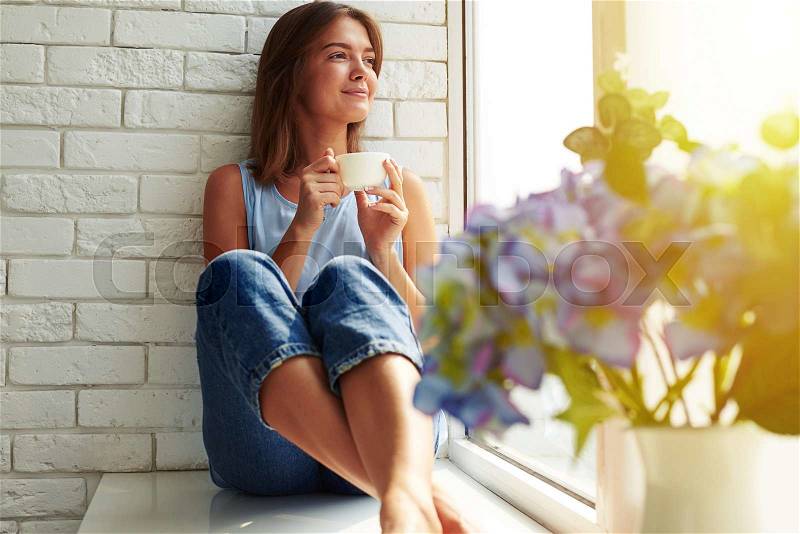 A well-pleased young girl enjoying an ideal day sitting in the window-sill. Wearing casual denim jeans and loose light-blue blouse, crossing legs and looking through the window, stock photo