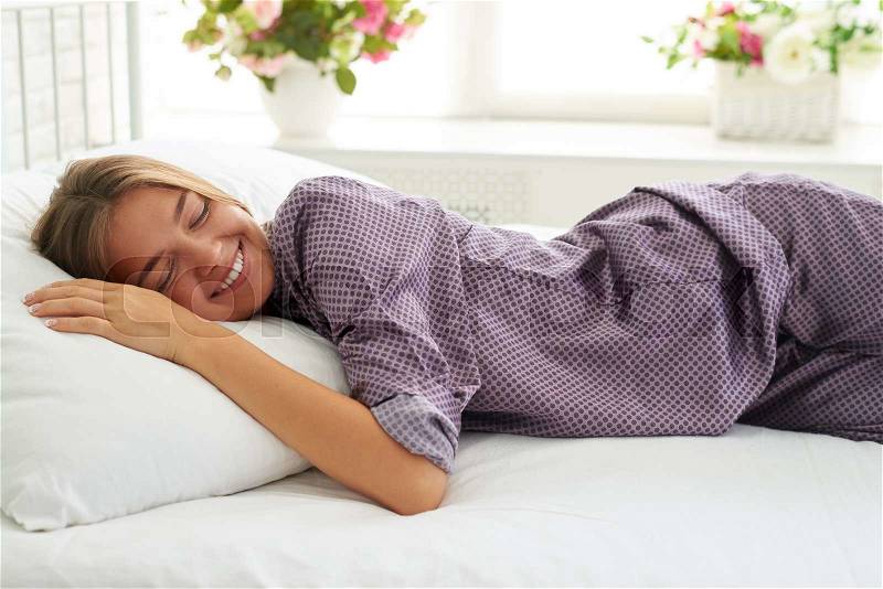 Portrait of smiling beautiful woman lying on the pillow on white bed in light bedroom with closed eyes, stock photo