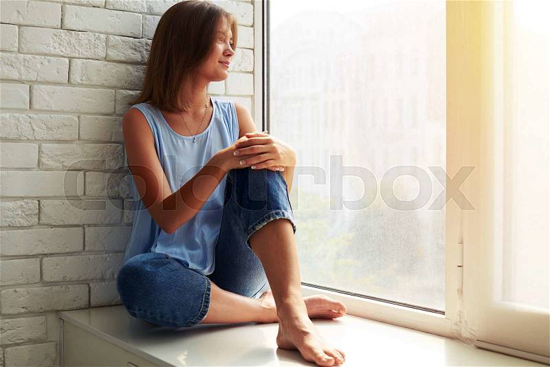 Thoughtful and dreaming girl looking out of the window while sitting on the window-sill, stock photo