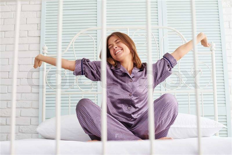 Young beautiful lady wakes up and does morning stretching sitting on bed with metal frame in light stylish bedroom, stock photo