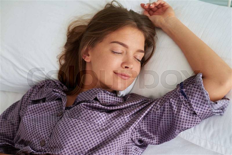 Young brown-haired woman in pajamas is sleeping peacefully on her back with one hand resting on pillow above her head, stock photo