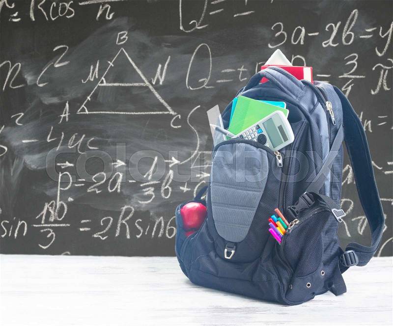 School backpack with supplies on white desktop, blackboard with math formulas in background, stock photo