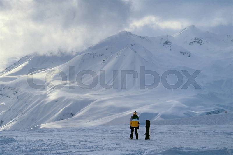 Away snowboarder stands and looks at mountains in winter, stock photo