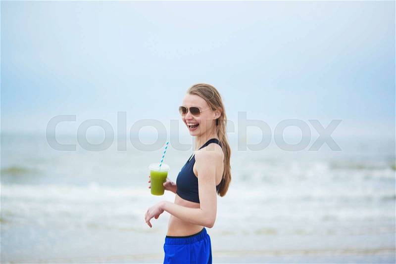 Healthy runner girl drinking green vegetable smoothie from plastic glass on running break. Young woman on beach cardio training taking a rest during workout. Healthy lifestyle, sport and diet concept, stock photo