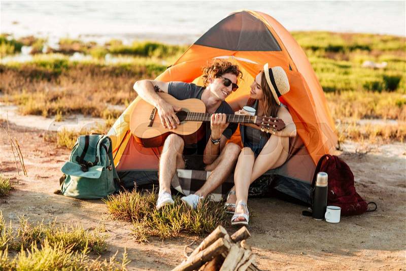 Portrait of a man playing guitar for his girlfriend sitting at the camping tent, stock photo