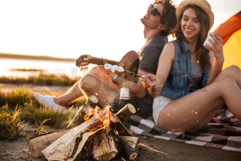 Cheerful beautiful young couple playing guitar and frying marshmallows on bonfire, stock photo