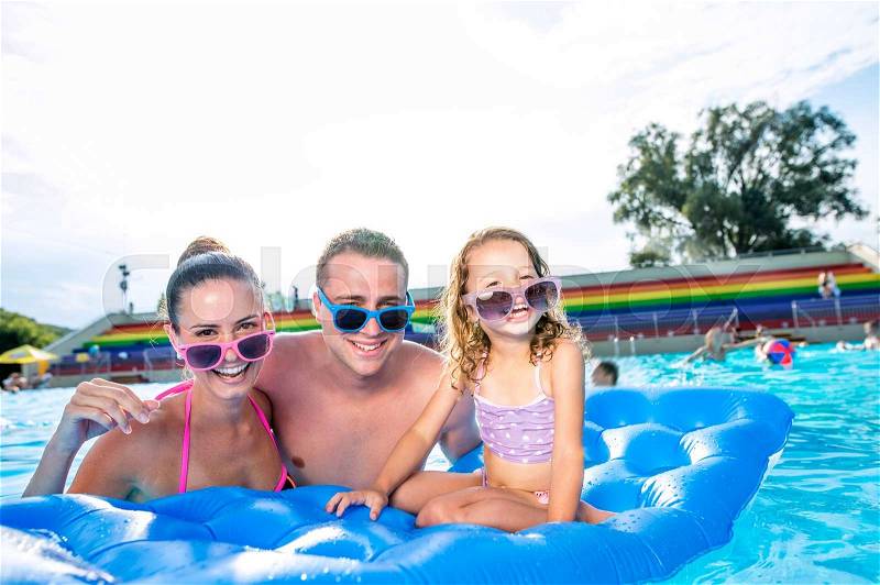 Young mother and father with their daughter in colorful sunglasses sitting on pool lilo in swimming pool in aqua park. Summer heat and water, stock photo