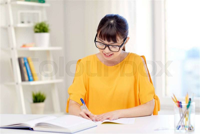 People, education, high school and learning concept - happy asian young woman student in glasses with book and notepad writing at home, stock photo