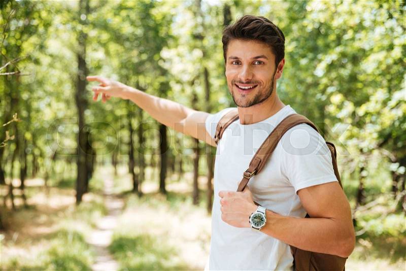 Cheerful young man with backpack standing and pointing away in forest, stock photo