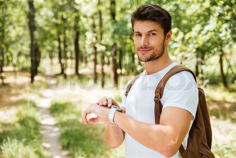 Smiling young man with backpack and watch standing in forest, stock photo