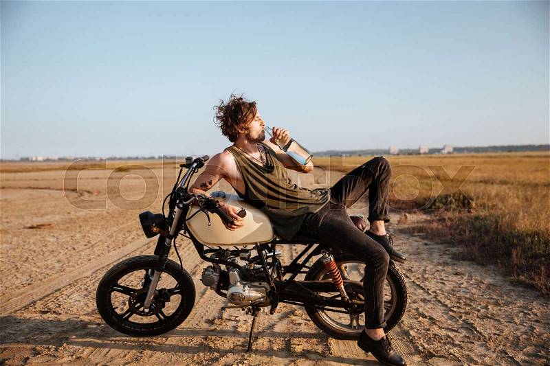 Young brutal man laying on his motorcycle in the desert and drinking water, stock photo