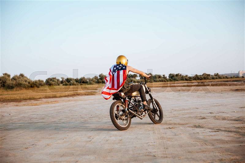 Young brutal man in golden helmet and american flag cape driving retro motorcycle, stock photo
