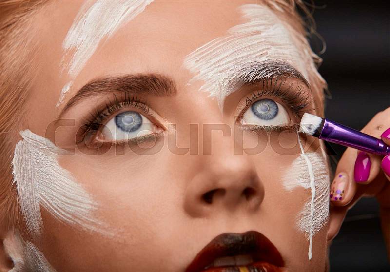 Process of bright art visage with white paint on girls the face. Beauty woman with creative make up and red plump lips. Brush makes a tear of white paint. Art visage on black background, stock photo