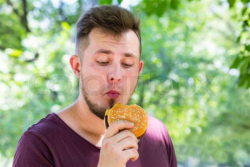 Emotional young guy eating a cheeseburger on the nature, stock photo