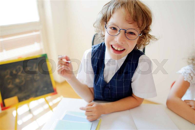 Funny little schoolboy in glasses sits at a school desk. It is a disciple of an elementary school. In a hand at boy ball pen. On a school desk copybooks lay. Near to a school desk a small blackboard, stock photo