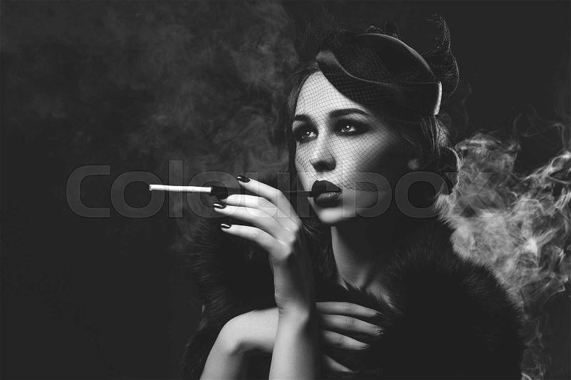 Beautiful young woman with smoky eyes and full red lips holding cigarette holder. Vintage head piece. Retro styling. Studio beauty shot over smoky background. Copy space. Monochrome, stock photo