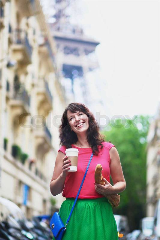 Cheerful young French woman walking with coffee to go and baguette (white bread) on a street of Paris, Eiffel tower in the background, stock photo