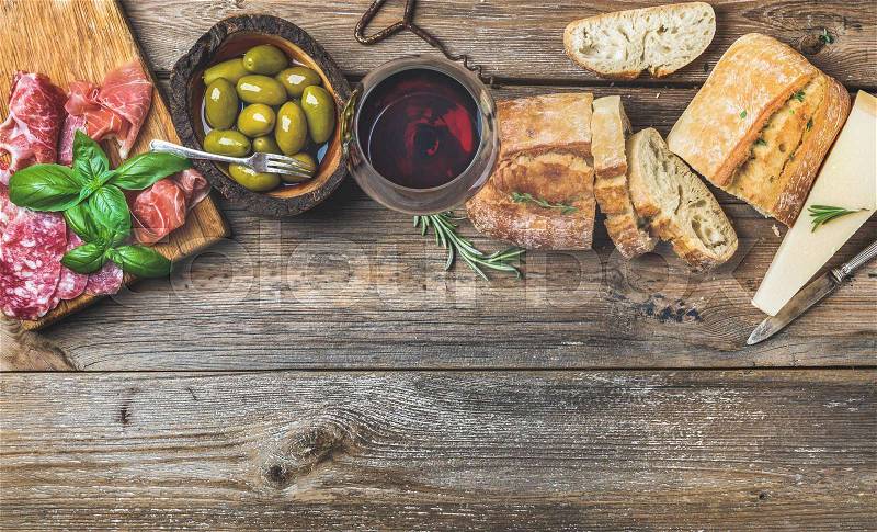 Wine snack set. Glass of red wine, green mediterranean olives, prosciutto, salami and aged cheese over rustic wooden background. Top view, copy space, stock photo