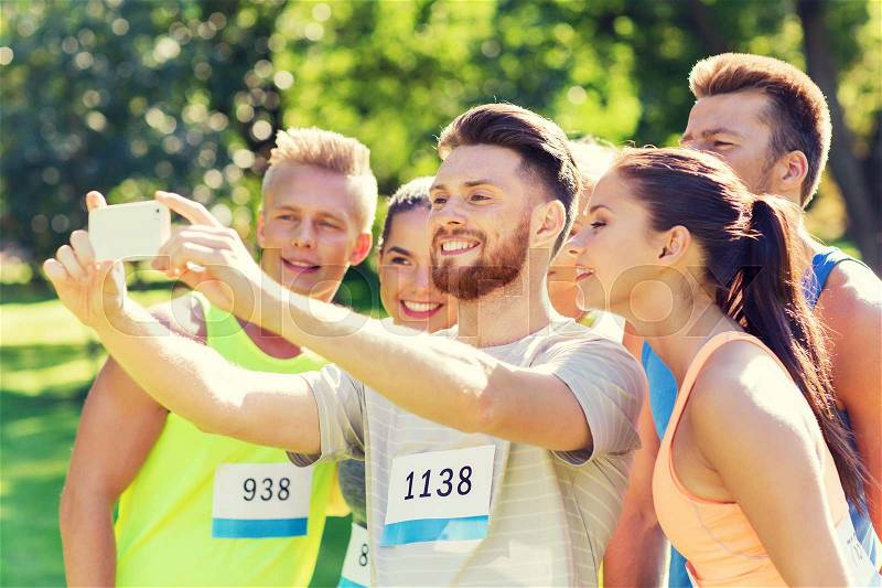 Fitness, sport, friendship, technology and healthy lifestyle concept - group of happy sportsmen friends with racing badge numbers taking selfie smartphone outdoors, stock photo