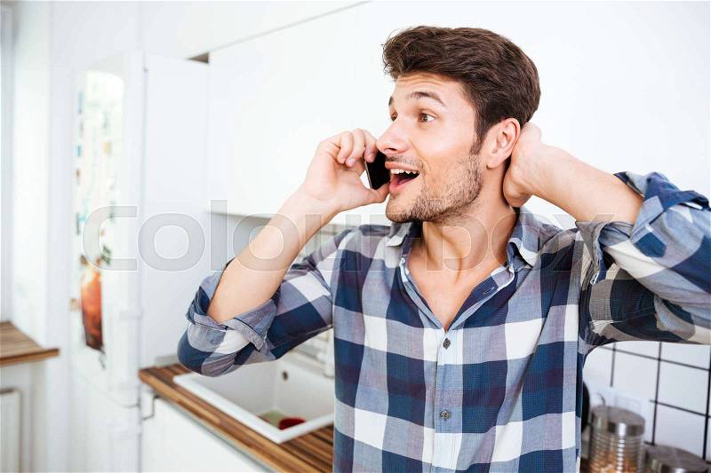 Happy amazed young man in checkered shirt talking on cell phone on the kitchen, stock photo
