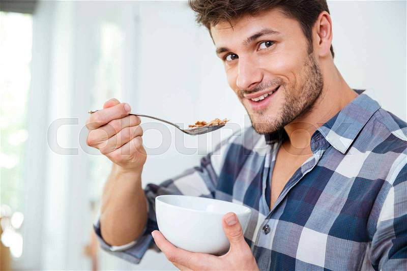 Portrait of happy young man in checkered shirt eating cereals with milk at home, stock photo