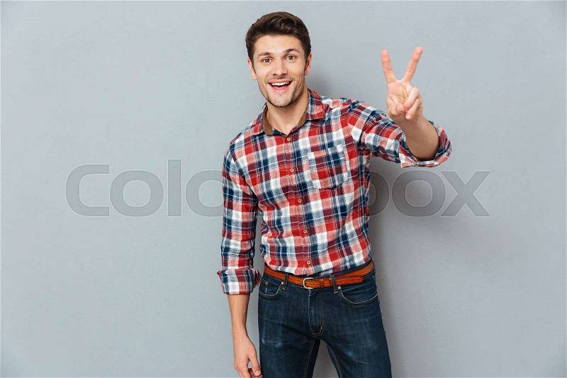 Handsome young man showing victory fingers sign over gray background, stock photo