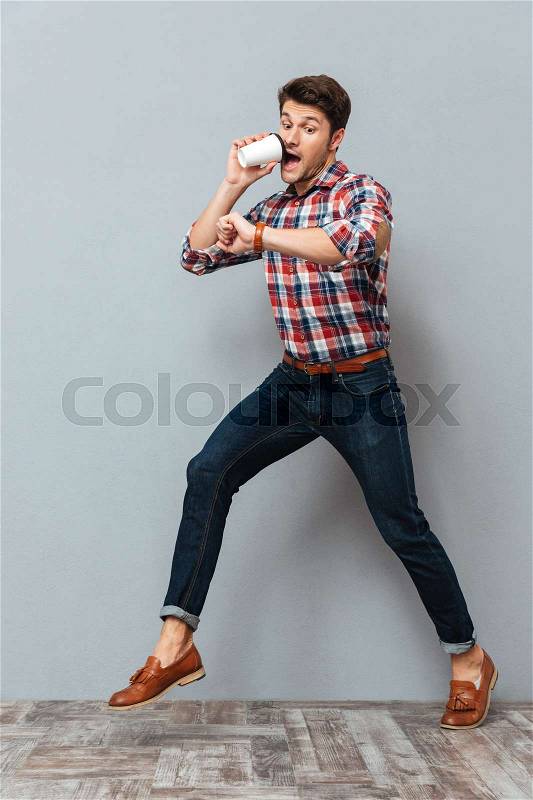 Shocked young man drinking takeaway coffee and running over grey background, stock photo