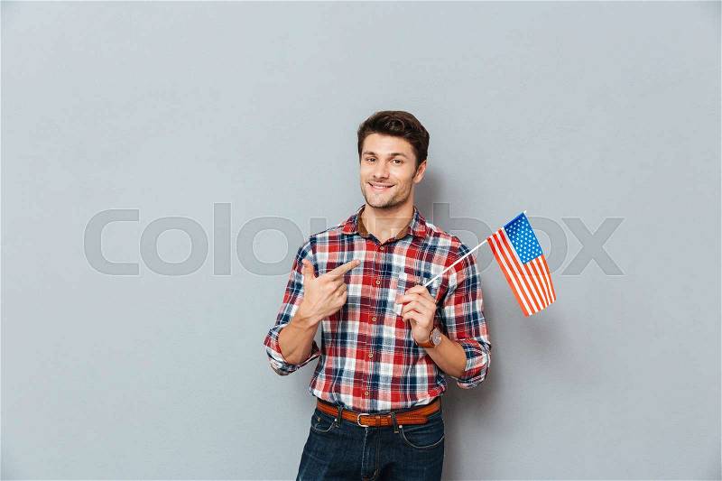 Smiling young man pointing on United States of America flag over grey background, stock photo
