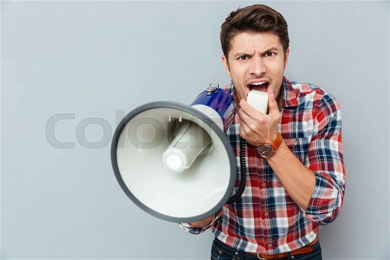 Mad crazy young man in plaid short shouting in megaphone over grey background, stock photo