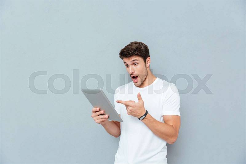 Surprised young man holding and pointing on tablet over grey background, stock photo