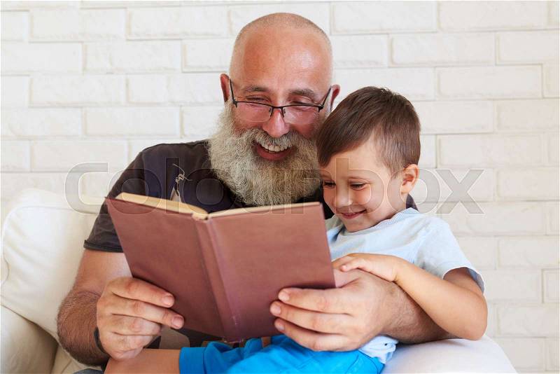 Aged man with white beard is reading a funny book to his grandson at home, stock photo
