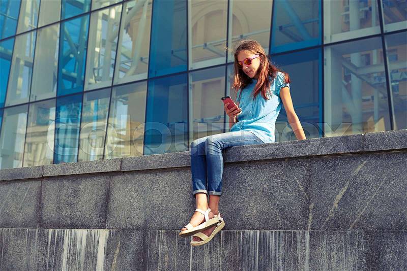 City lifestyle stylish youth using a smartphone sitting against the business on a sunny summer day, stock photo