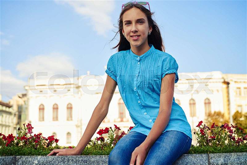 Close-up of teenage girl in bright blue blouse sitting on the flowerbed on street and posing at the camera, stock photo