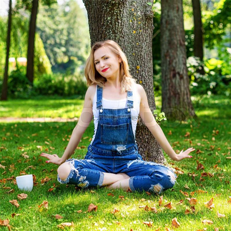 Happy blonde woman enjoying nature sitting on green grass with cup of coffee. Freedom concept, stock photo