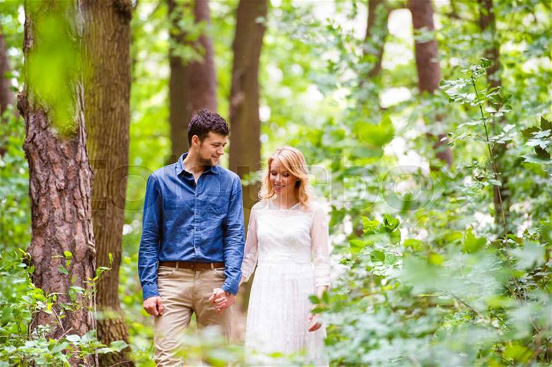 Beautiful young wedding couple on a walk outside in green forest, holding hands. Bride in white dress and groom in denim shirt, stock photo