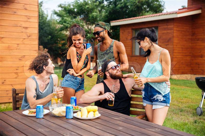 Happy group of teenage friends sitting at table drinking beer and eating snacks, stock photo