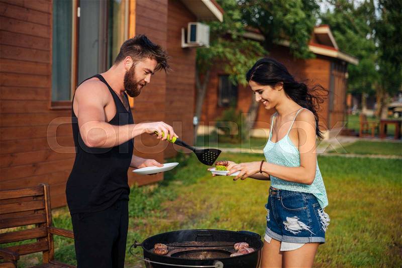 Happy young couple standing and cooking meet on barbeque grill outdoors, stock photo