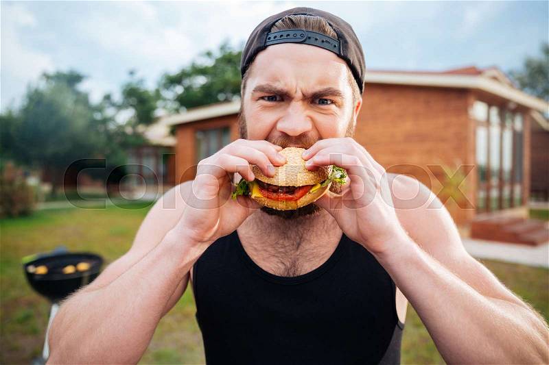 Young bearded hipster guy eating delicious burger outdoors, stock photo