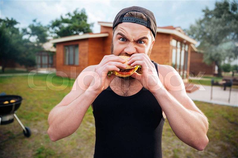 Young bearded hipster guy biting delicious burger outdoors, stock photo