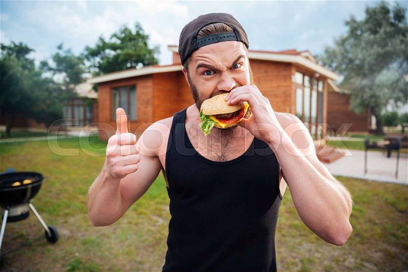 Young bearded hipster guy biting delicious burger and showing thumbs up outdoors, stock photo