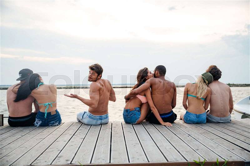 Sad confused young man sitting between happy hugging couples on pier, stock photo