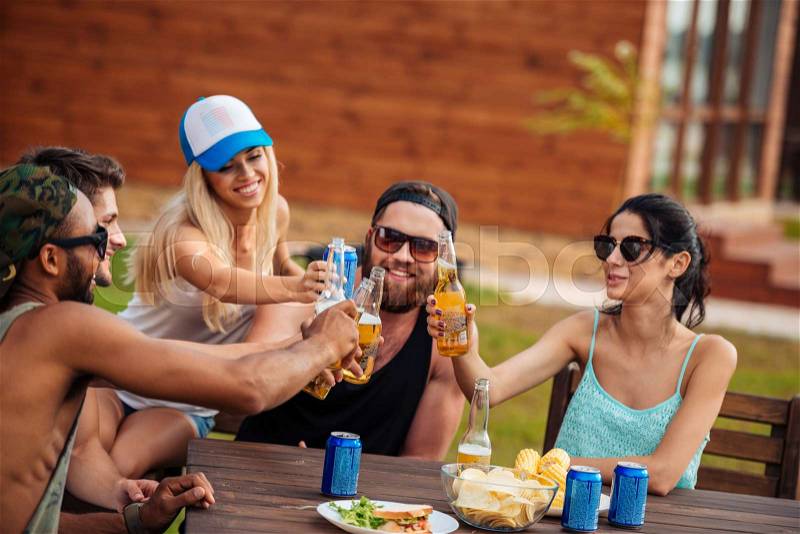 Happy group of teenage friends sitting at table drinking beer and having fun outdoors, stock photo