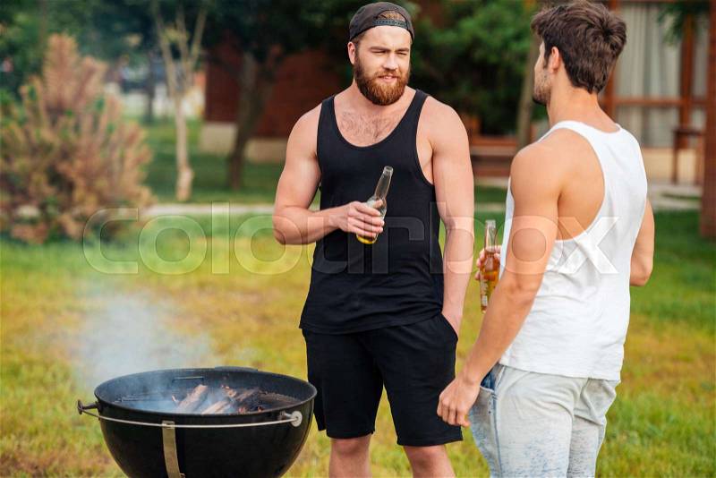 Two men holding a beer bottle while preparing barbecue grill in park zone, stock photo