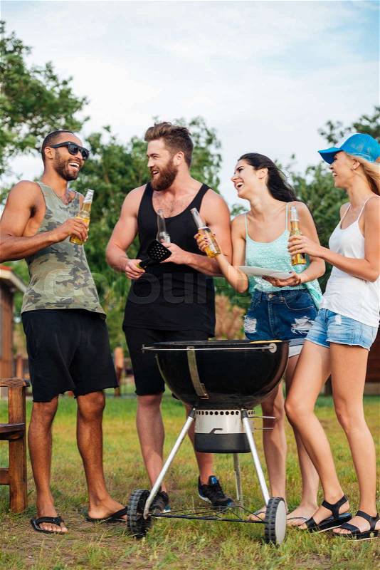 Group of cheerful young friends drinking beer and frying meet on barbeque grill outdoors, stock photo