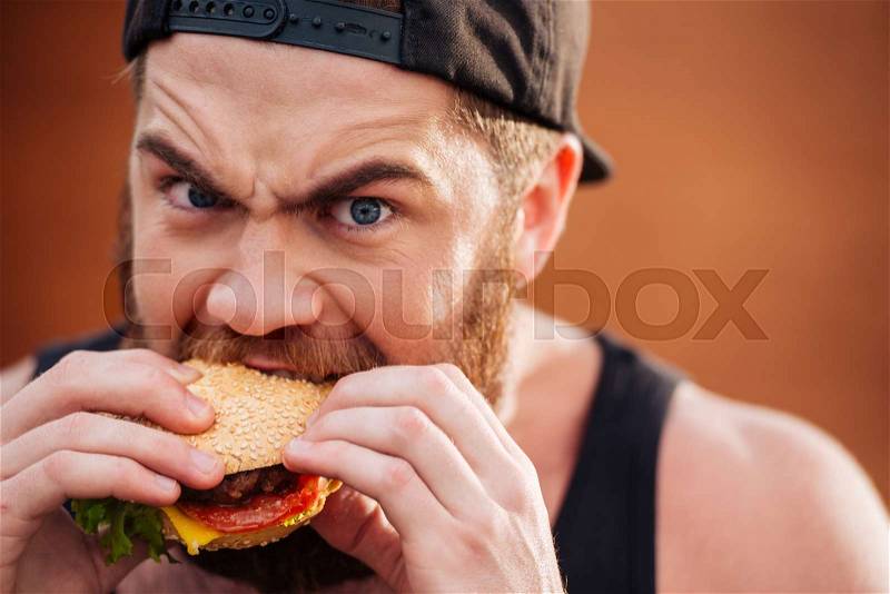 Angry irritated young man in cap eating hamburger outdoors, stock photo