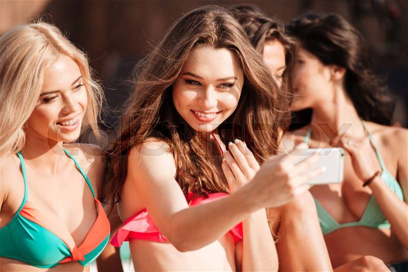 Four pretty young girls in bikinis making selfie with phone at the swimming pool, stock photo