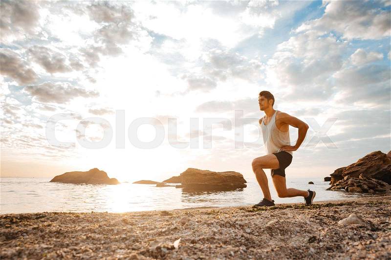 Young healthy man athlete doing squats at the beach at sunset, stock photo