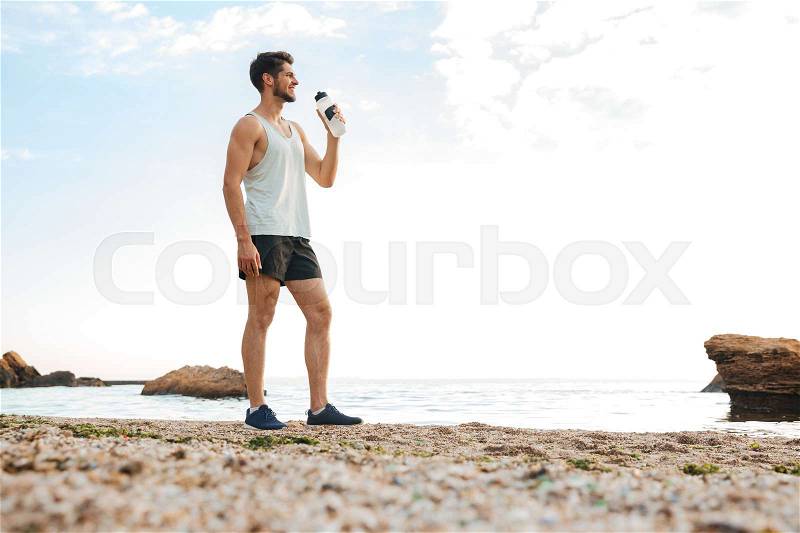 Young sporty man athlete resting after jogging at the beach and drinking water, stock photo