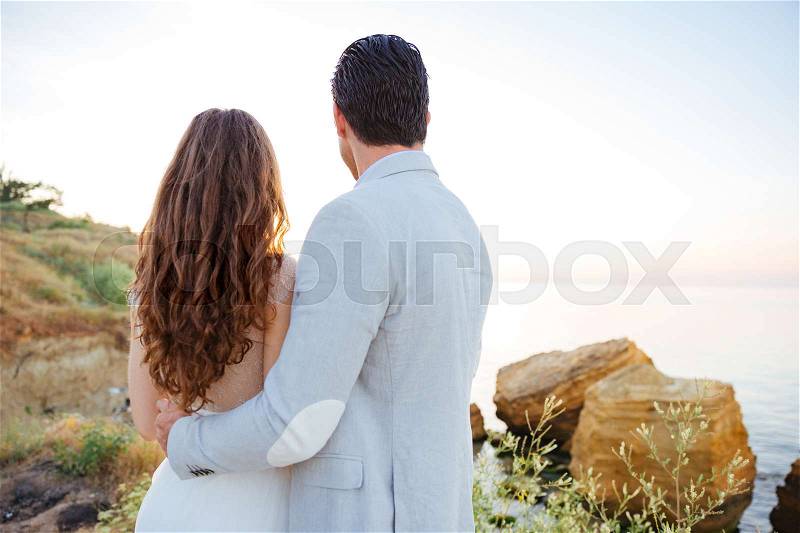 Back view of a romantic married couple hugging on the beach at sunset, stock photo