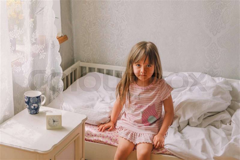Little girl sitting on the bed. Little girl is wearing pajamas anjd sitting on the bed in the bedroom, next to the table is an alarm clock and a cup of water, stock photo